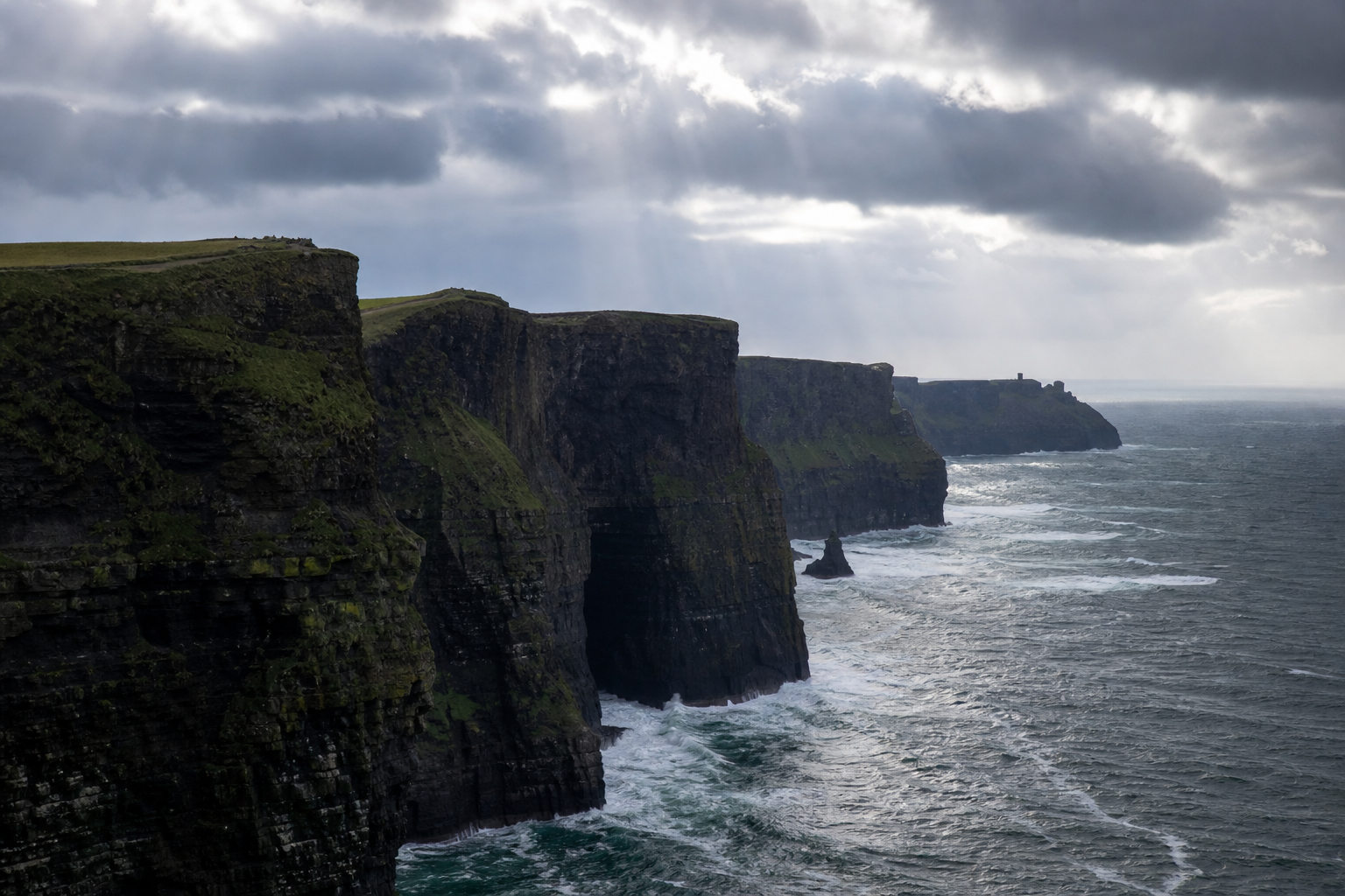 The Cliffs of Moher Ireland, towering green-topped sea cliffs dropping into the Atlantic, crashing white waves below, dramatic cloudy Irish sky, real photograph, landscape photography