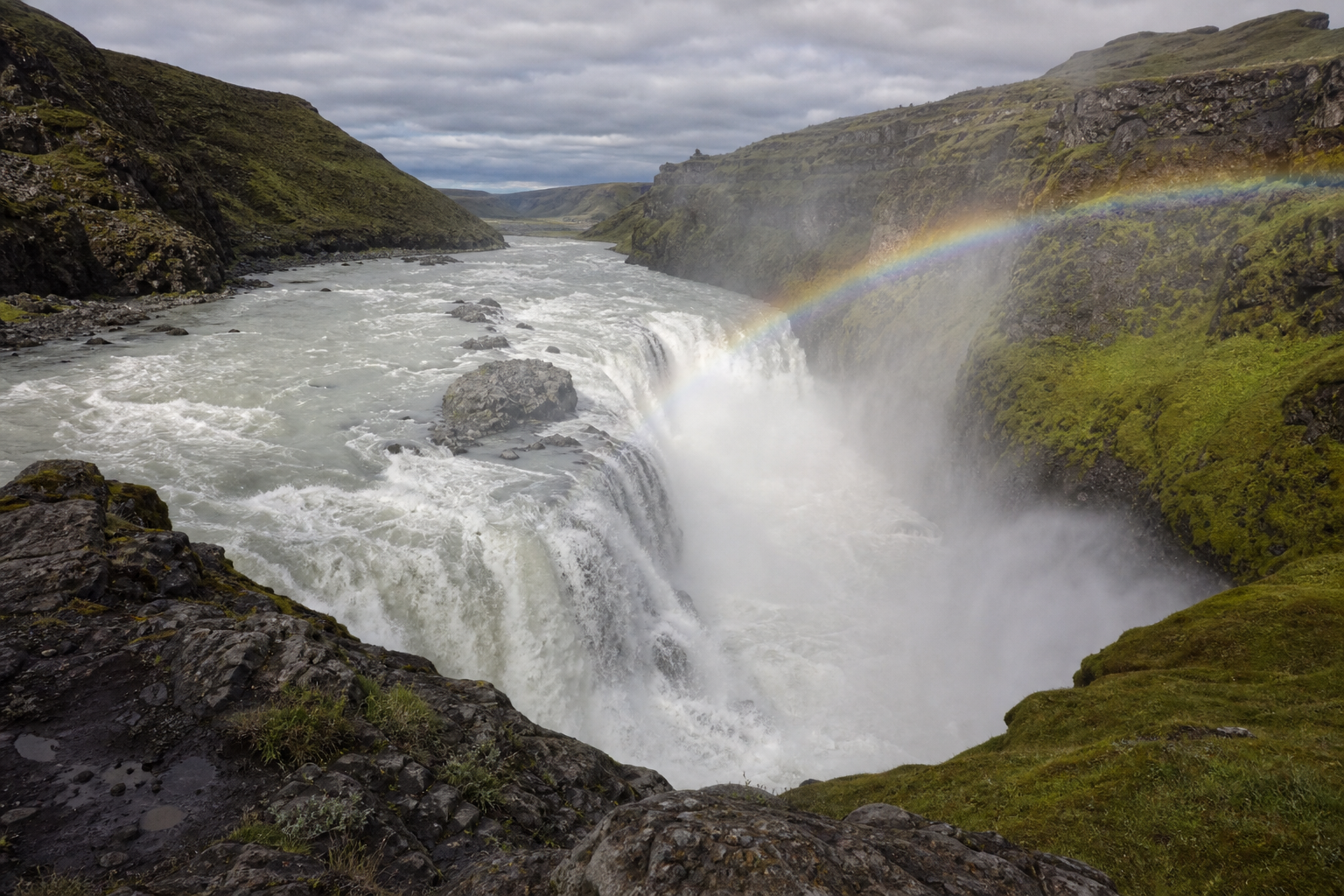 A powerful Iceland waterfall cascading into a misty gorge, black volcanic rocks and lush green moss, rainbow forming in the spray, real photograph, landscape photography